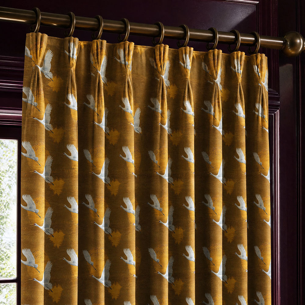 Luxury velvet fabric featuring elegant flying cranes on a rich ochre background with painterly cloud textures styled as statement curtains within a dramatic interior
