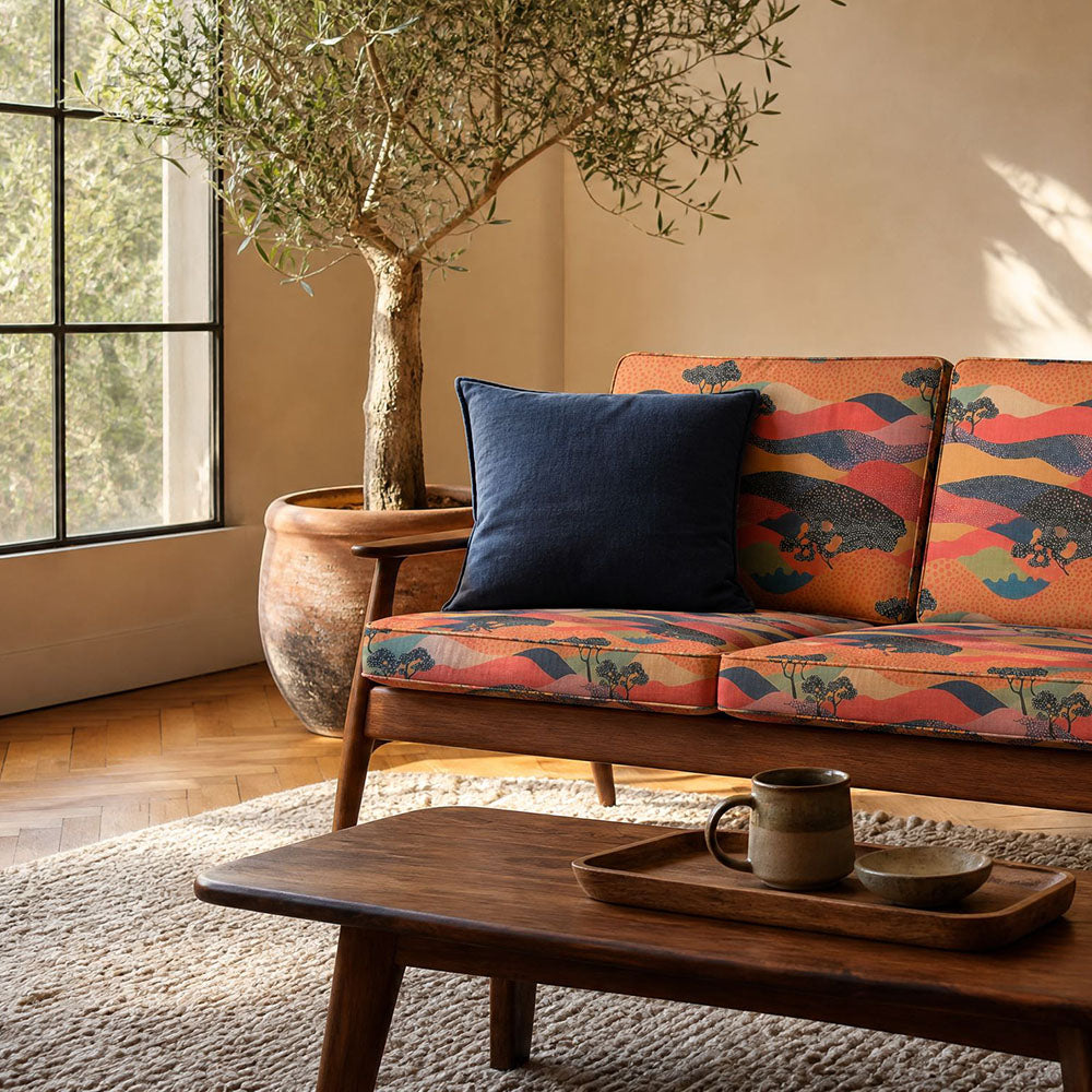 Japanese upholstery fabric featuring layered mountain landscapes, rolling hills and silhouetted trees in coral, terracotta, navy and gold tones upholstered onto a contemporary sofa